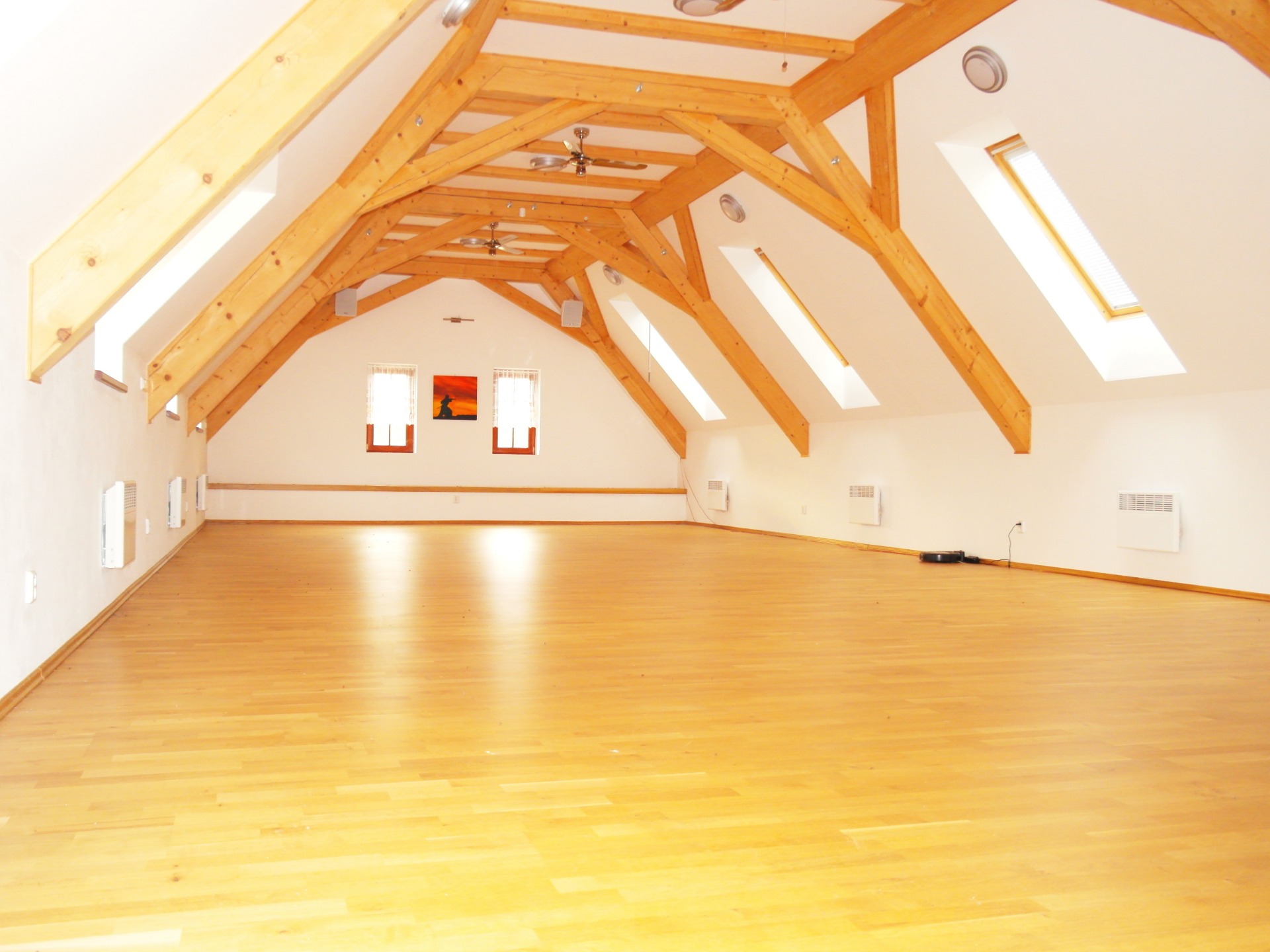 Prostorná místnost s dřevěnými trámy v podkroví, parketovou podlahou, stropními ventilátory a okny.