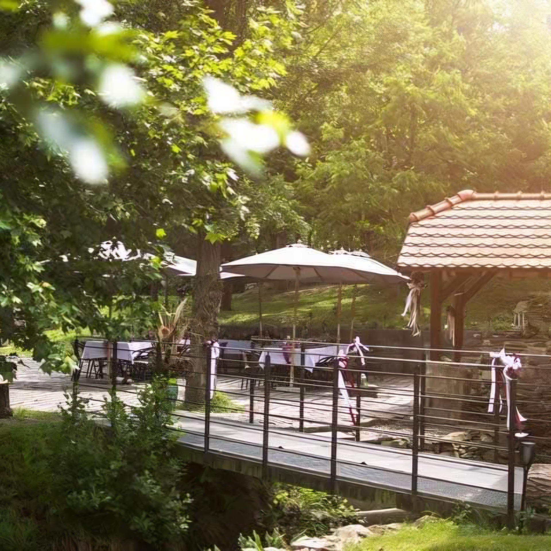 Zahradní terasa s bílým slunečníkem, dřevěným pergolovým stínem a zelenými stromy v teplém slunečním světle.